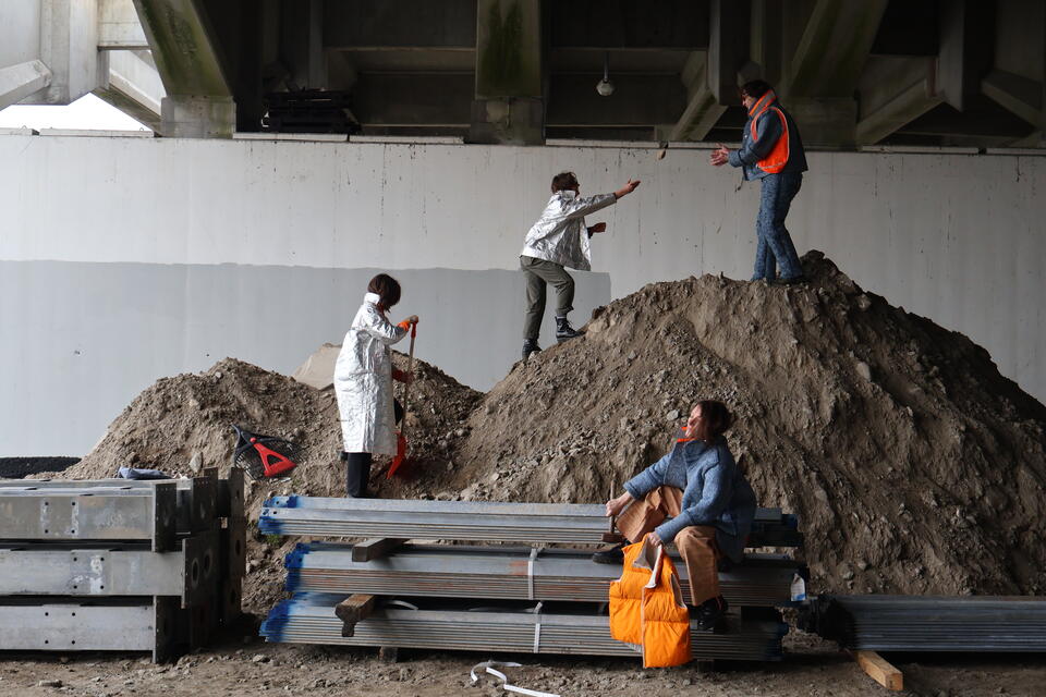 Photograph of four people wearing the following garments:100% Recycled Denim Suit and Jacket, two Tyvek Jackets, cork chaps, and an orange nylon vest stuffed with recycled denim pulp, in a construction site under an overpass.