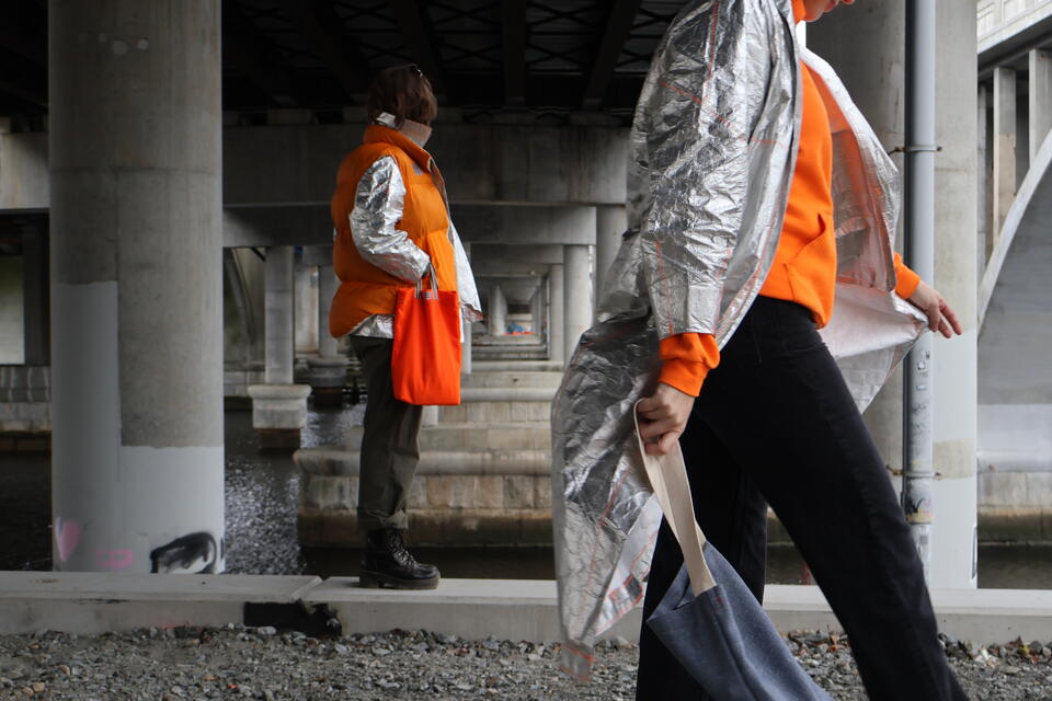 Photograph of two people wearing the Tyvek Trench and Tyvek Coat layered with a Shoddy Vest under an overpass, holding bags.