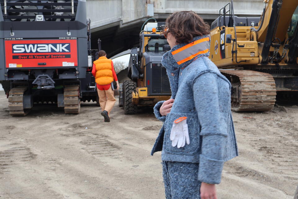 Photograph of two people, one in the foreground wearing the Denim Insulation Suit, the other person is walking away, wearing the orange Shoddy Vest and Cork Chaps.