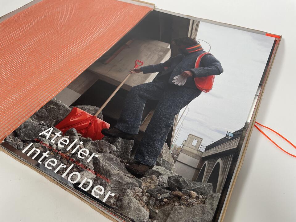 Photograph of the collection's lookbook opened to the cover page, a photograph of a person in the denim insulation suit climbing the mountain of rubble with an orange shovel. Title of the page reads "Atelier Interloper"