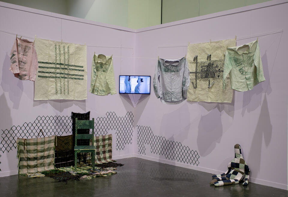 Image of printed clothes hanging from clothesline, video performance, woodcut prints, carved chair, woven fabrics, and stuffed doll