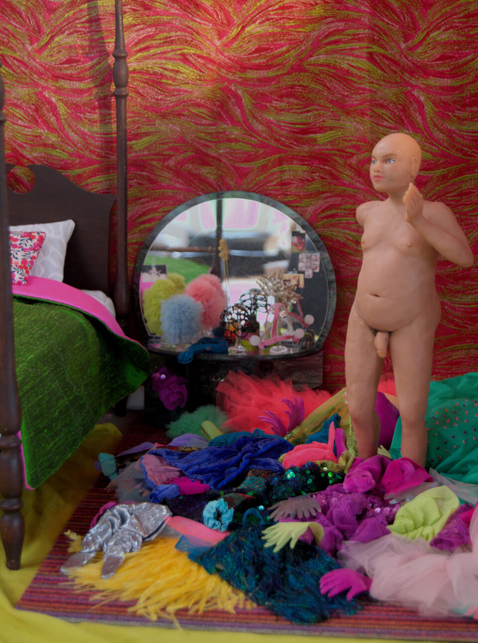 A sculpted nude figure stands in a bedroom, the floor covered with elaborate, brightly-colored clothes. A mirrored vanity has ruffled headdresses on glass wigstands, a post bed is covered with a green and pink silk duvet.
