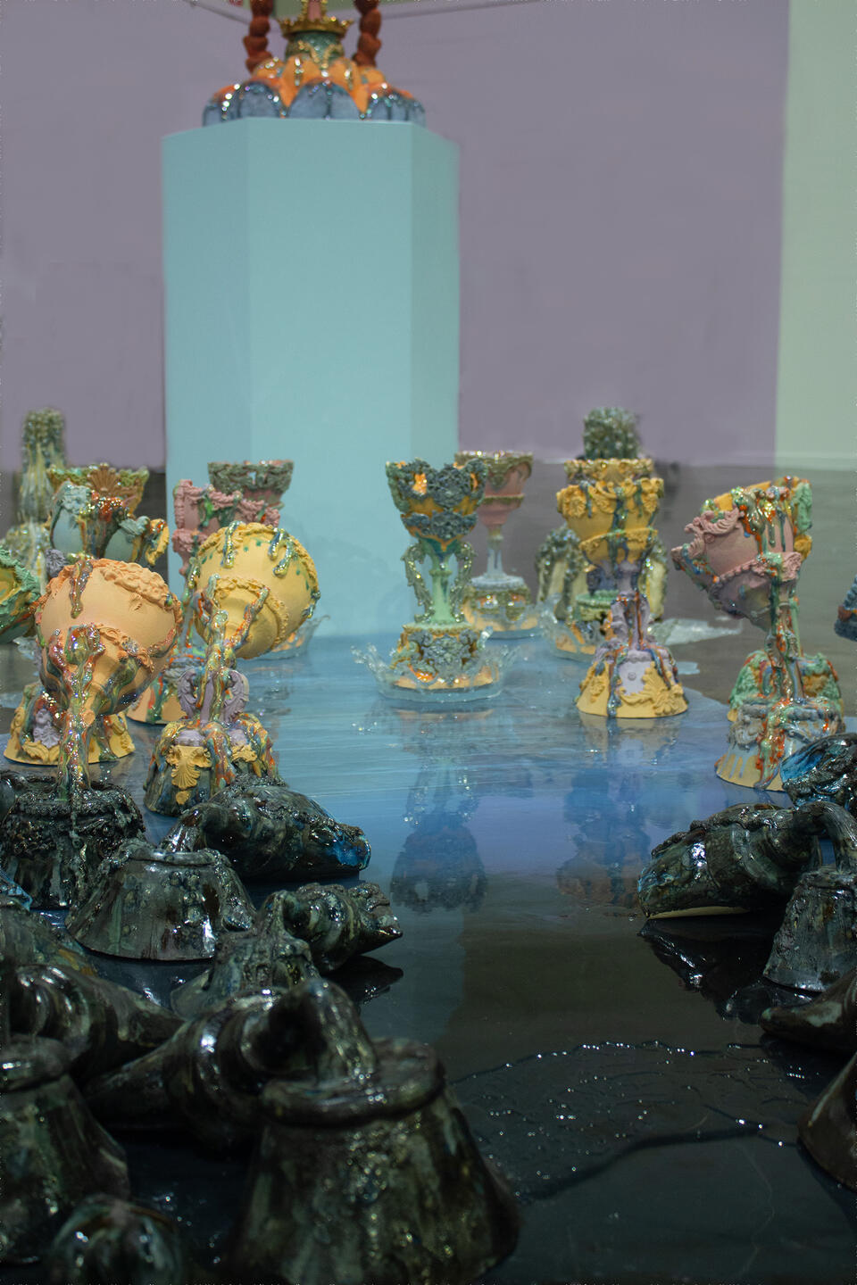 Colorful ceramic vessels arranged in clusters on a reflective surface, with a central pedestal holding an elaborate piece. The holy grails are gradually changing colors.   