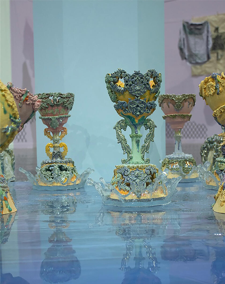 Close-up of intricately decorated the Holy grail with dripping glazed surfaces and covered with pastel colors, standing on a glossy blue reflective surface which is water.