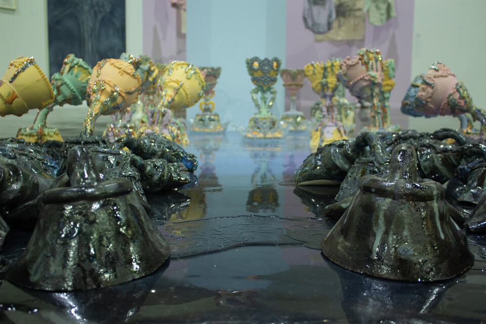 Foreground features dark, glossy ceramic forms with a rugged texture to express water, leading to pastel-colored, ornate vessels in the background on a reflective surface. 