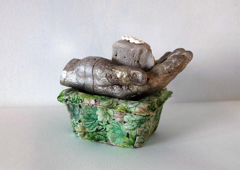 a stack of objects; a white mold of three molar teeth, an ocean animal vertebrae, a pewter hand that is crushing a blackberry plastic container that is covered in green leaf patterned fabric