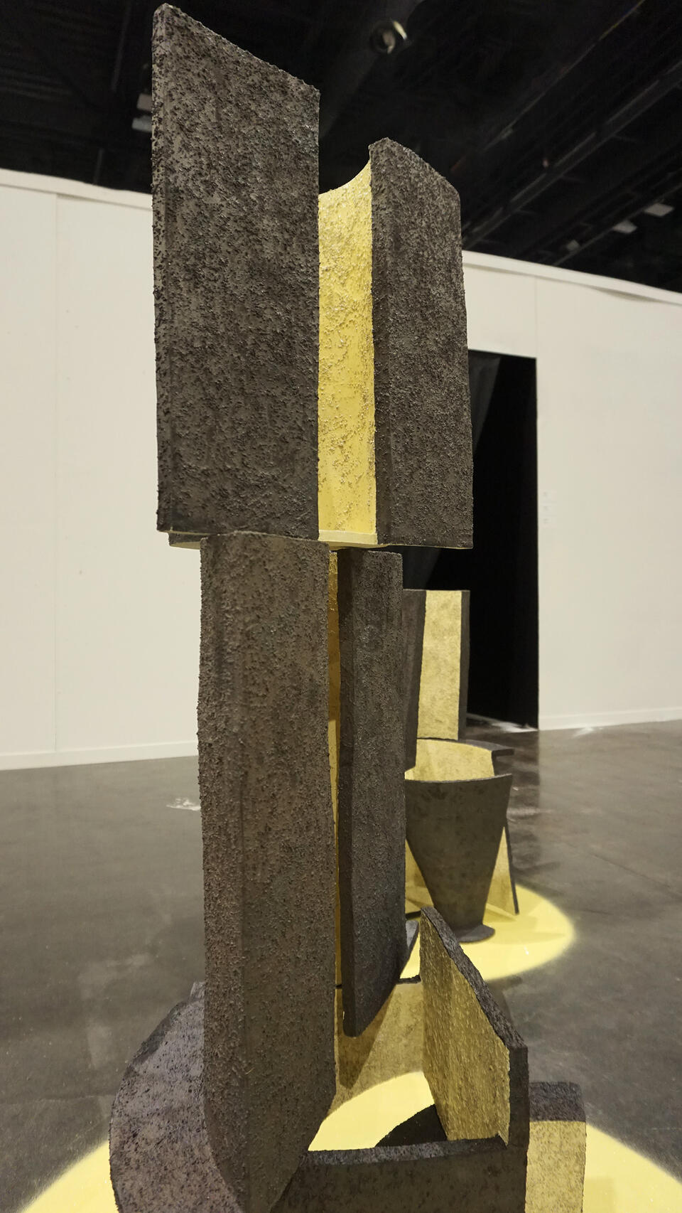 Close-up of a textured, geometric ceramic sculpture with vertical panels in black and yellow, displayed on a yellow circular base. The background shows additional sculptures with similar designs.
