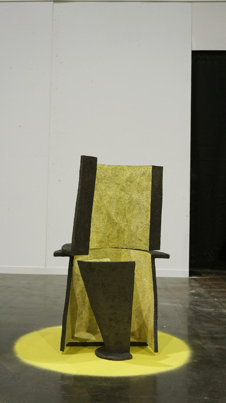 A textured, geometric ceramic chair with a tall back and angular seat, predominantly yellow with black accents, is placed on a yellow circular base in a minimalist gallery space.