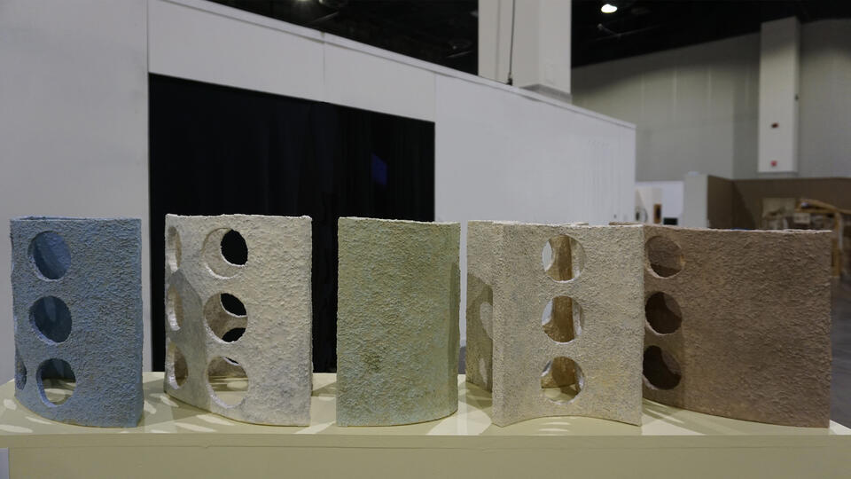 A row of textured, crescent-shaped ceramic sculptures with circular cutouts, arranged in gradient of earth tones and pastels. The foreground shows beige, transitioning to green and blue in the background.