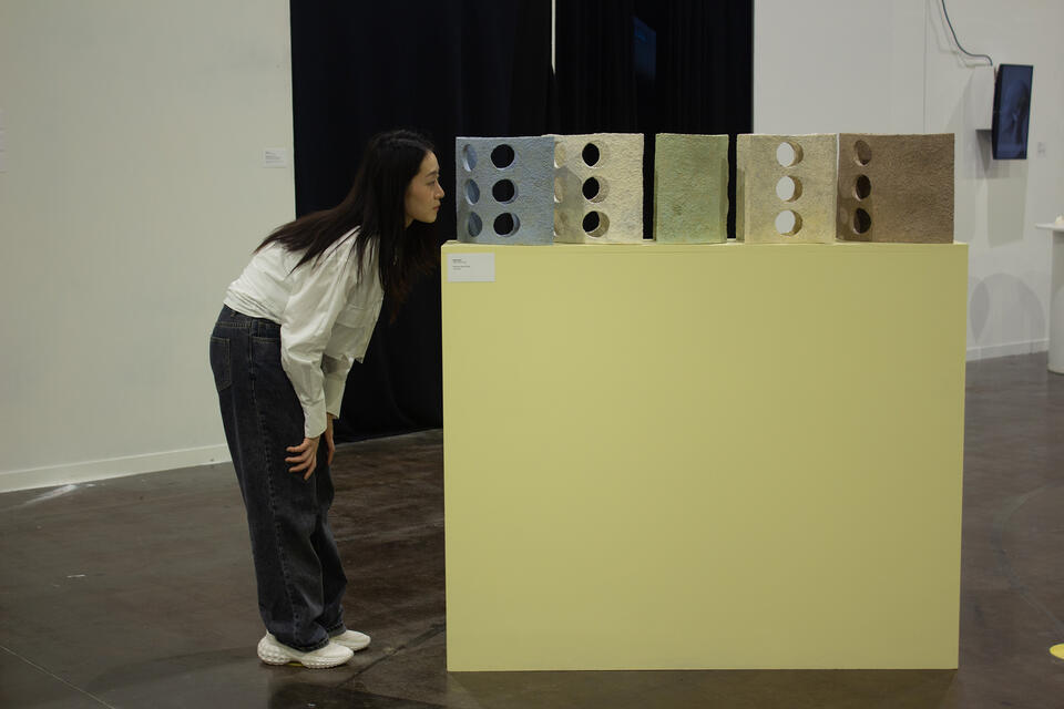 A woman in a white blouse and dark jeans leans forward to closely examine a series of textured, crescent-shaped ceramic sculptures with circular cutouts, displayed on a light yellow pedestal.