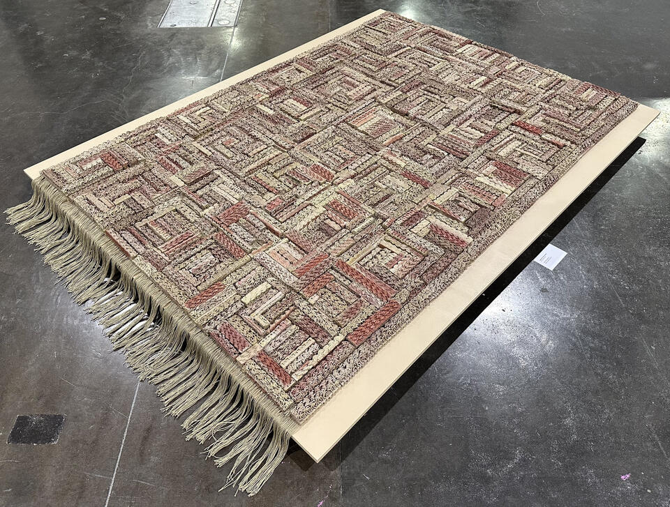 3D-printed ceramic (Egyptian paste) piece with intricate patterns in shades of red, beige, and brown. The first row is made of yarn with hanging threads. The piece is displayed on a wooden platform, placed on a polished concrete floor.