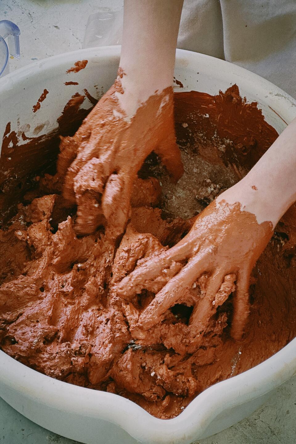 Hands mixing reddish Egyptian Paste in a white bowl, creating a textured, earthy mixture. The scene captures the tactile and immersive process of preparing ceramic material.