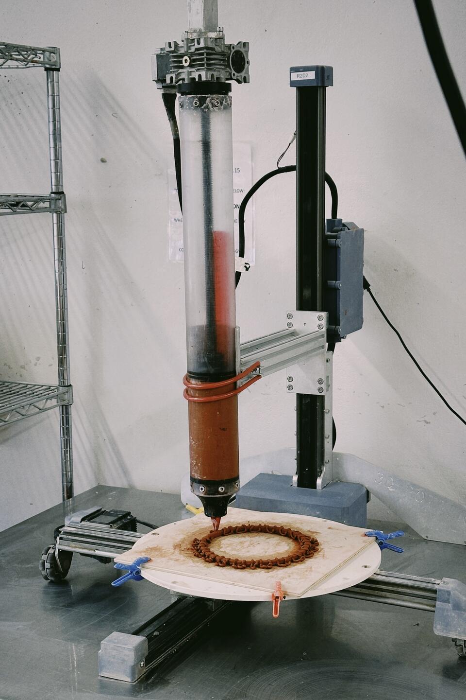 3D printer extruding a circular clay pattern on a rotating platform, with the printer labeled 'R2D2' in a workshop setting.
