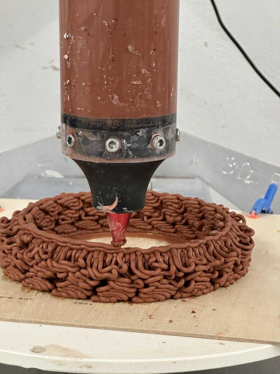 3D printer nozzle depositing intricate, layered clay structure onto a wooden platform, capturing the detailed formation process.