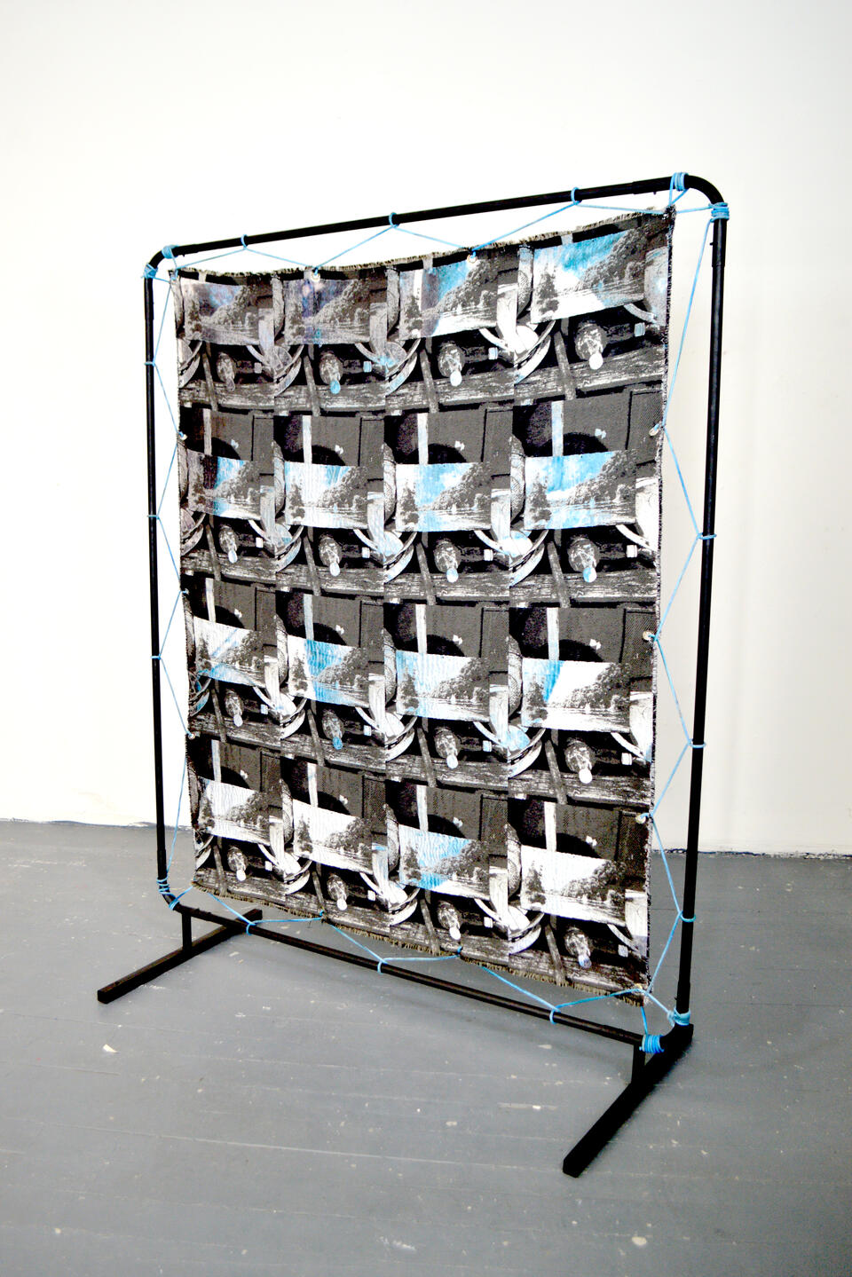 Large black steel frame standing in a studio facing a blank wall with a black and white photographic weaving depicting an image repeat. The weaving is stretched using a blue rope.