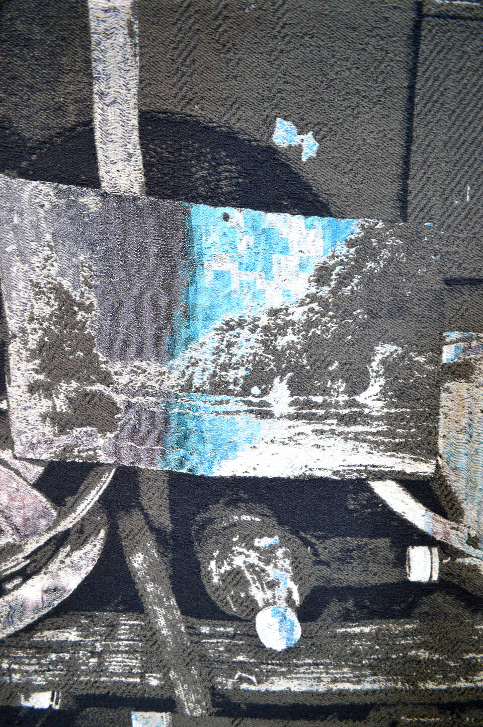 An image depicting a close up of a weaving. The close up shows one repeat of a larger work with a landscape and a plastic bottle in a greyscale color palette.