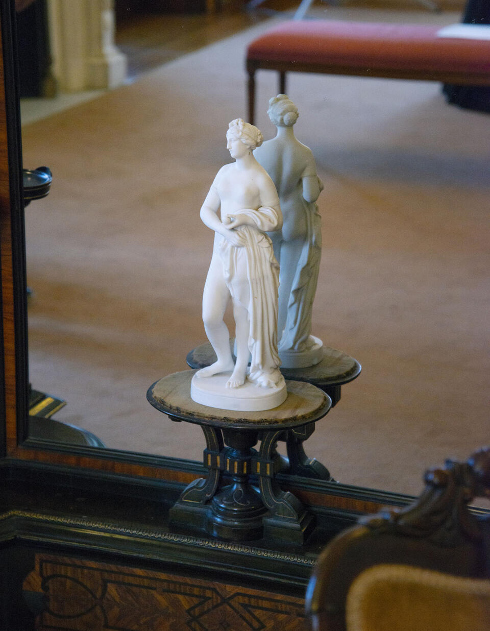 A marble statue of the Greco-roman goddess, Venus, posed against a mirrored background. Reflected in the mirror is the carpet of an interior room.  