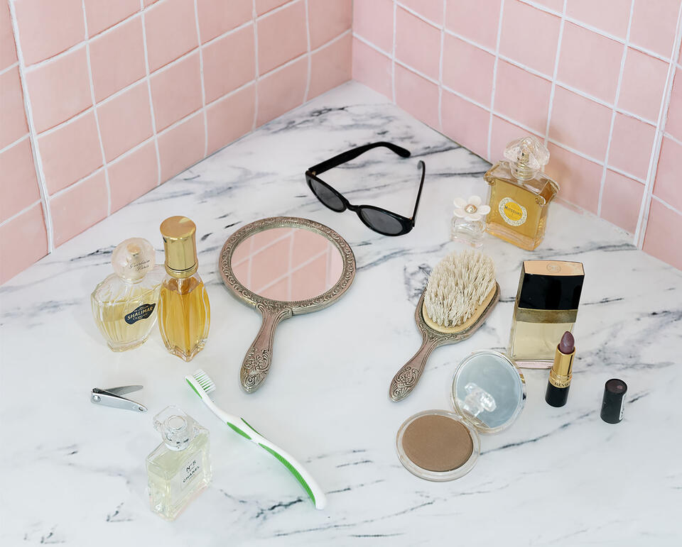 A still life of a marble countertop against pink ceramic tiles with perfume bottles, a hairbrush, compact mirror, sunglasses, and makeup.