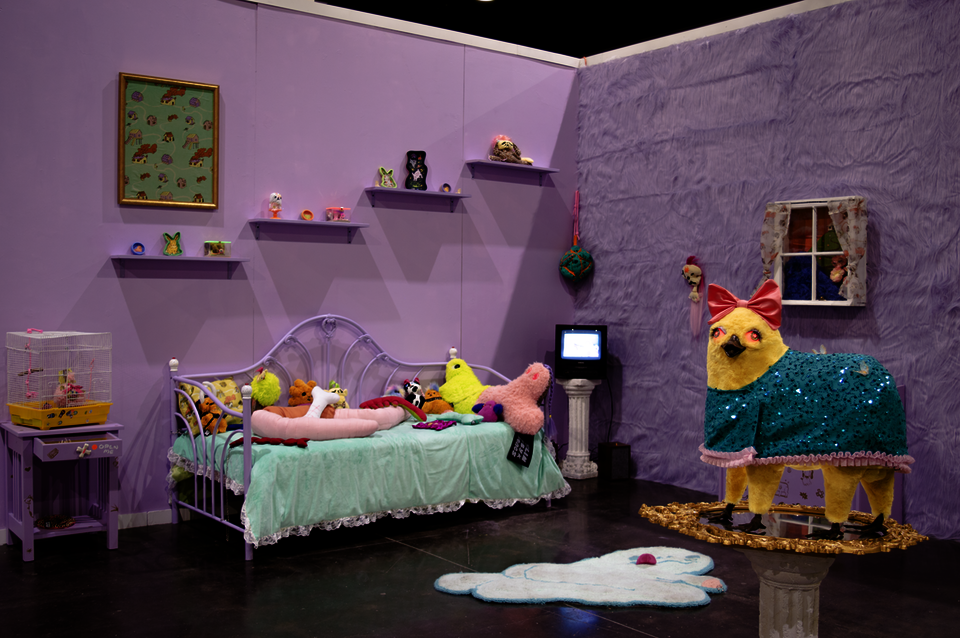 Photograph of an installation of a fantasy bedroom.  There is a bed covered with toys, a TV in the corner playing a reel of chaotic videos, and sculpture of a duck/horse chimera.  