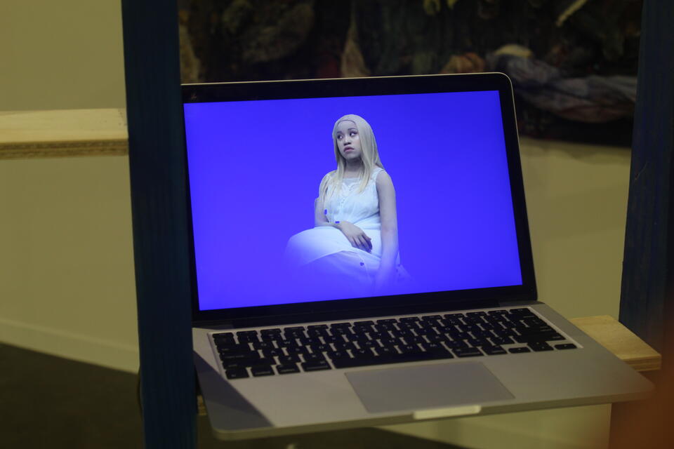 The image shows the artist in a video still, wearing pale face paint, a blond wig, and a pale blue nightgown, against a fluorescent blue background. The video plays on a MacBook Air 2019.