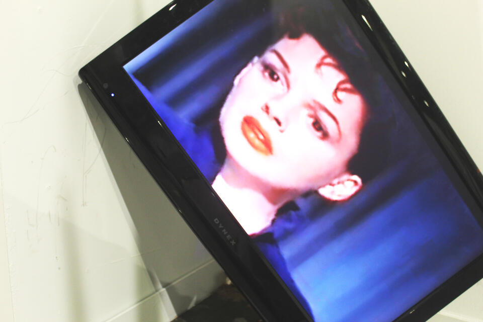 A large rectangular monitor propped in the corner of a white room shows a closeup of Judy Garland's face, looking startled and concerned.