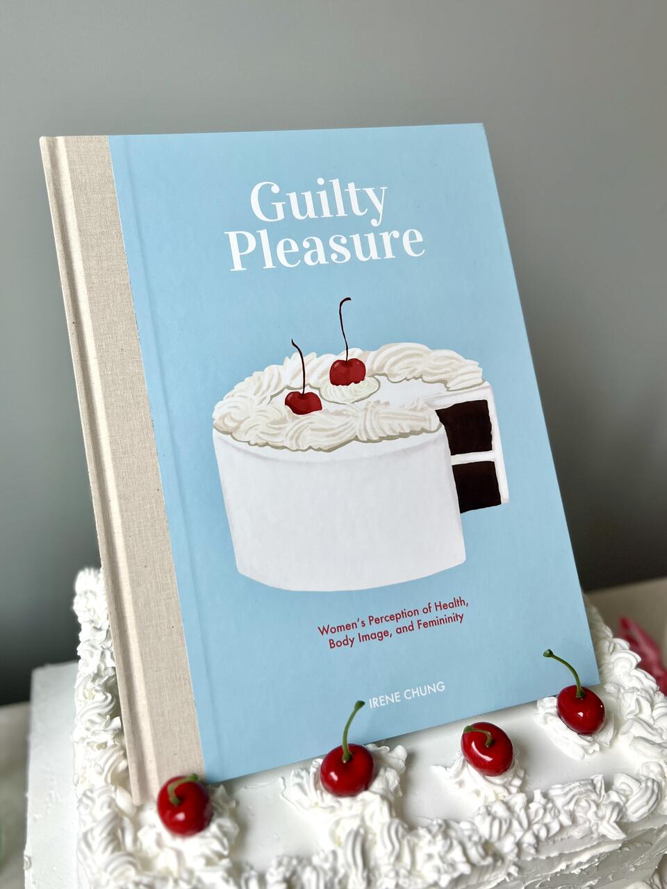 an illustrated book, titled Guilty Pleasure, sitting on a cake with cherries on top 