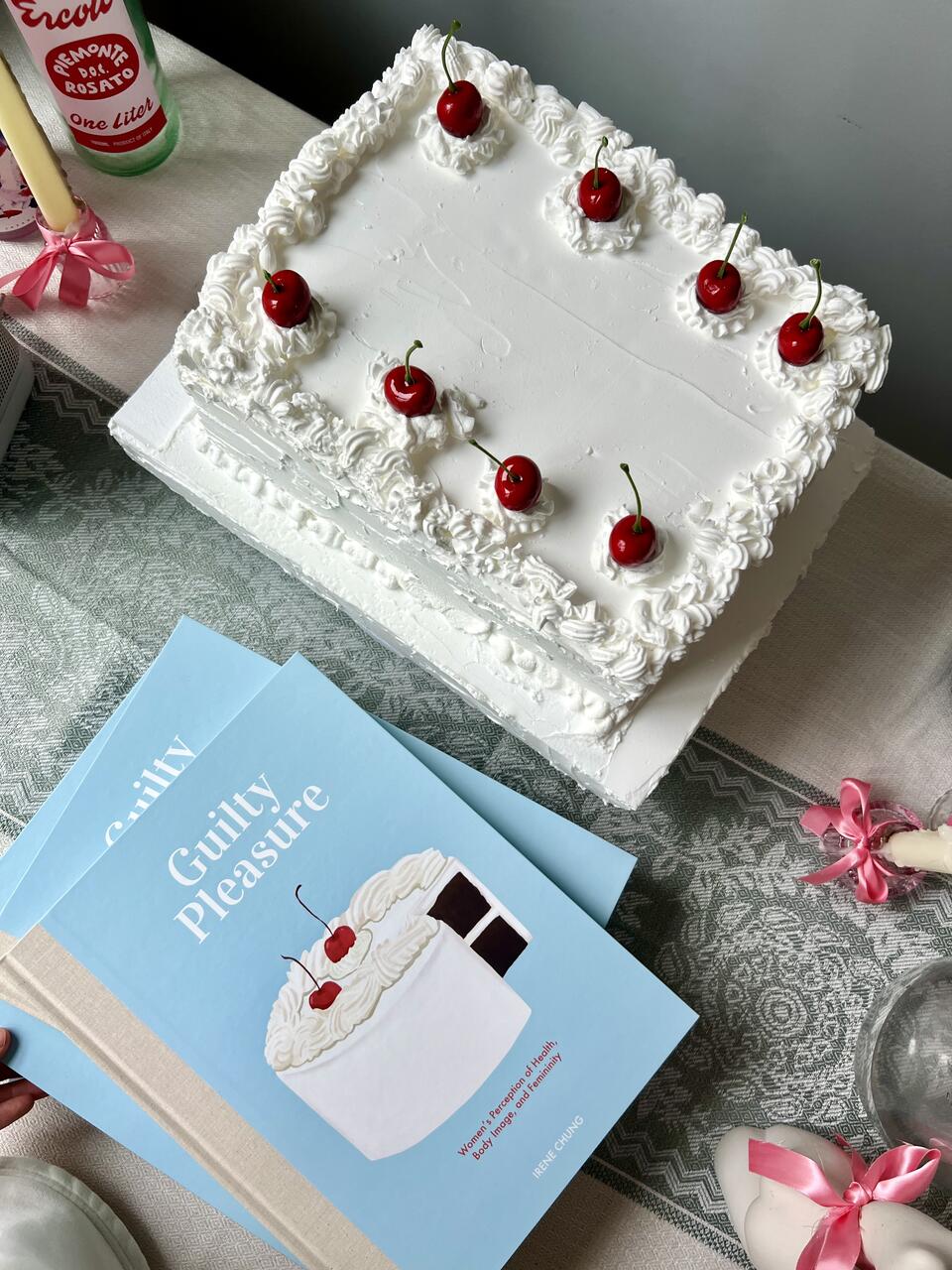 photo of illustrated books next to a white-cream cake with cherries on top, on the surface of tablecloth