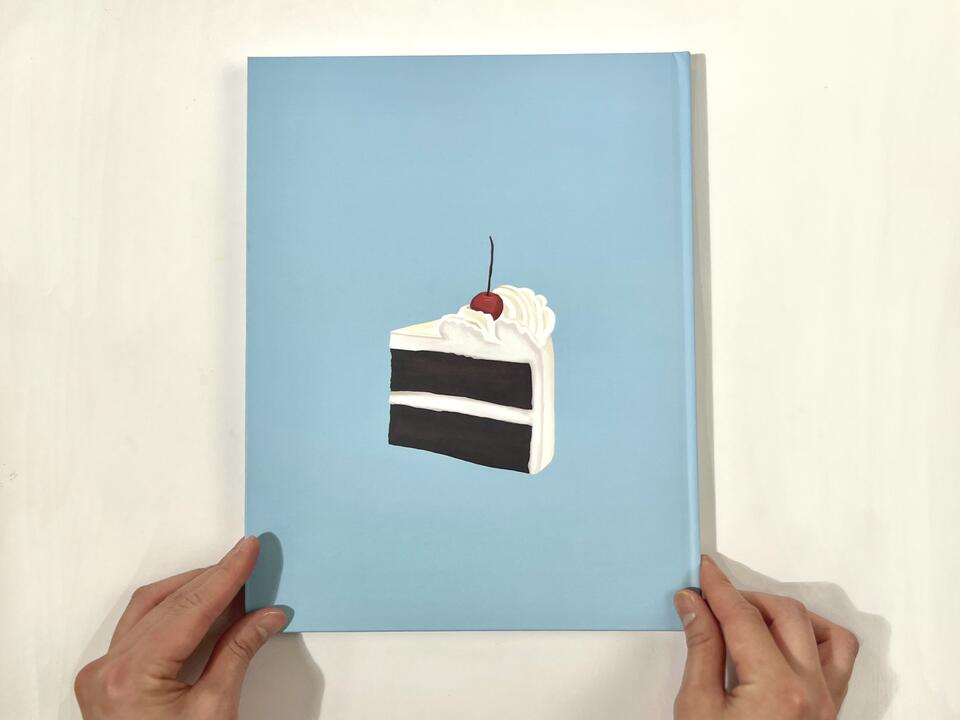 A photograph of the book cover, which features a piece of creamy white cake with a cherry on top, sitting on a baby-blue background