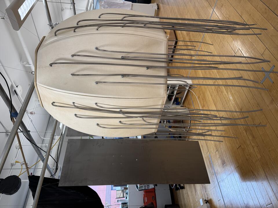 Barrel-shaped goatskin structure with hanging metal springs, surrounded by steel sheets and lit by overhead projection.