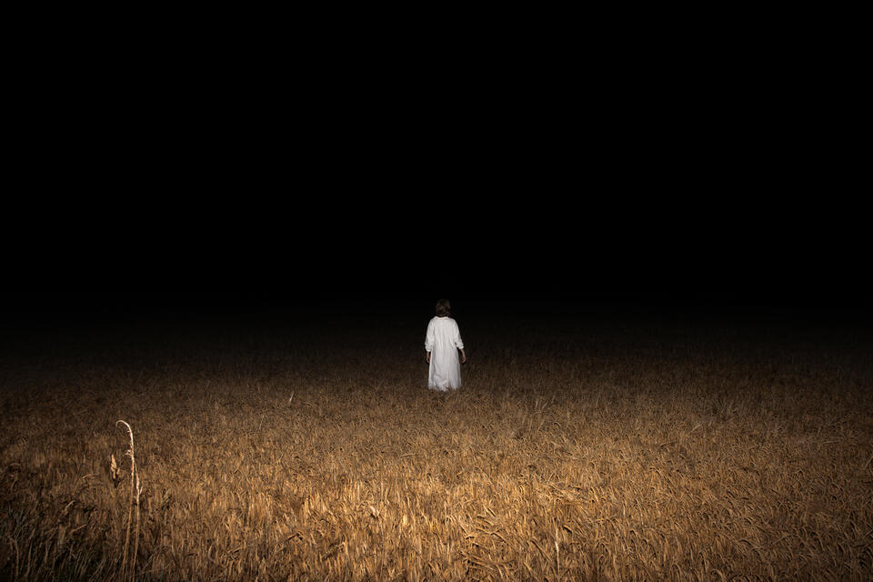 A figure in a white Sankta Lucia gown stands in a wheat field. She faces the dark, void of the night.