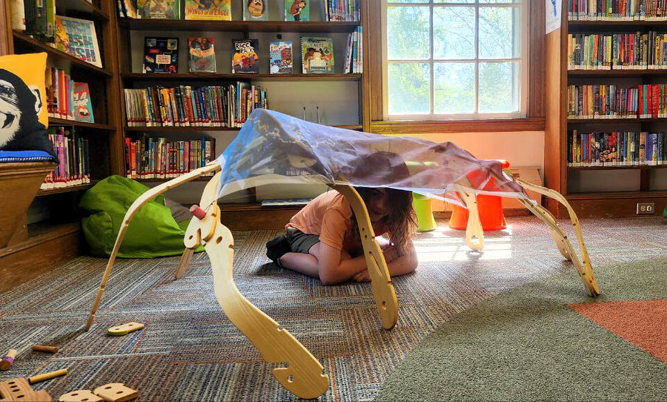 Child sitting inside a wooden toy structure covered in printed fabric with ash tree leaves, seeds, bark, and twigs