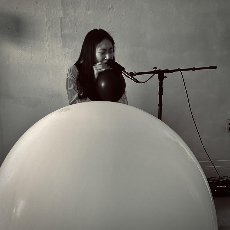 A performer creates balloon noise with a microphone, processed through a computer, and played back via speakers.