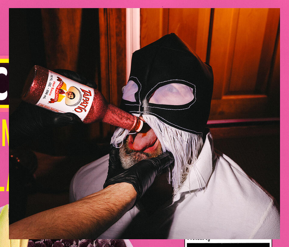 A masked man in a lucha libre hood with white fringe is force fed pink glittered Tapatío on his tongue by an unknown hand
