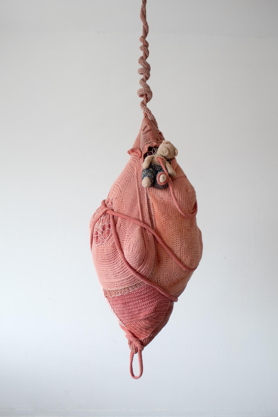 pink, pupa-shaped crochet sculpture with tapering ends, hanging by a twisted cord; a small crochet teddy bear emerges.