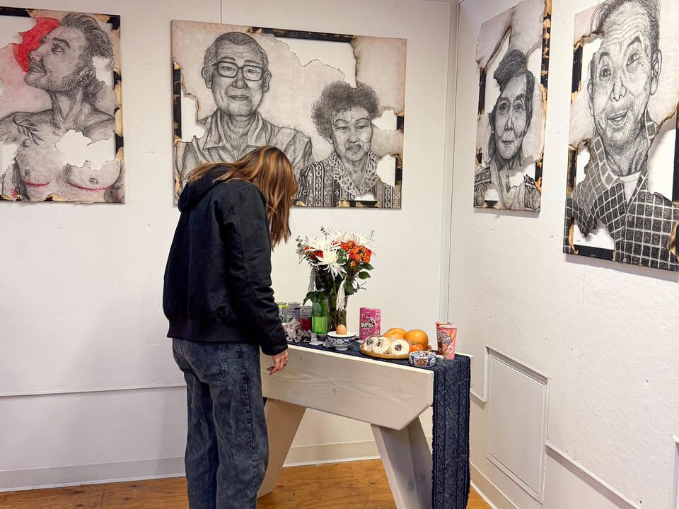 A person stands and examines an altar holding food and fresh flowers. On the walls are partially burned charcoal portraits.